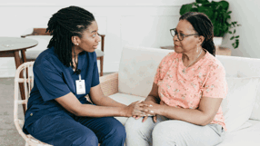 Seated health care worker holding hands with elderly lady sitting on sofa